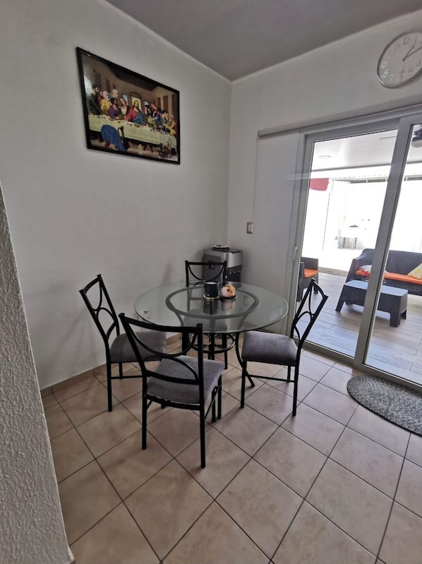 Uma área de refeições com uma mesa de vidro e quatro cadeiras, um quadro na parede e um relógio acima da porta.