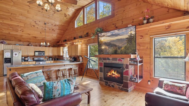 A cozy cabin interior with a stone fireplace, wooden furniture, and large windows.