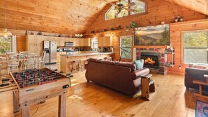 A cozy wooden cabin interior with a foosball table, a fireplace, and a large window overlooking a forest.