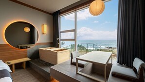 Habitación de hotel moderna con un ventanal grande que ofrece vista al mar, un espejo redondo, un banco de madera, una pequeña mesa y un sofá.