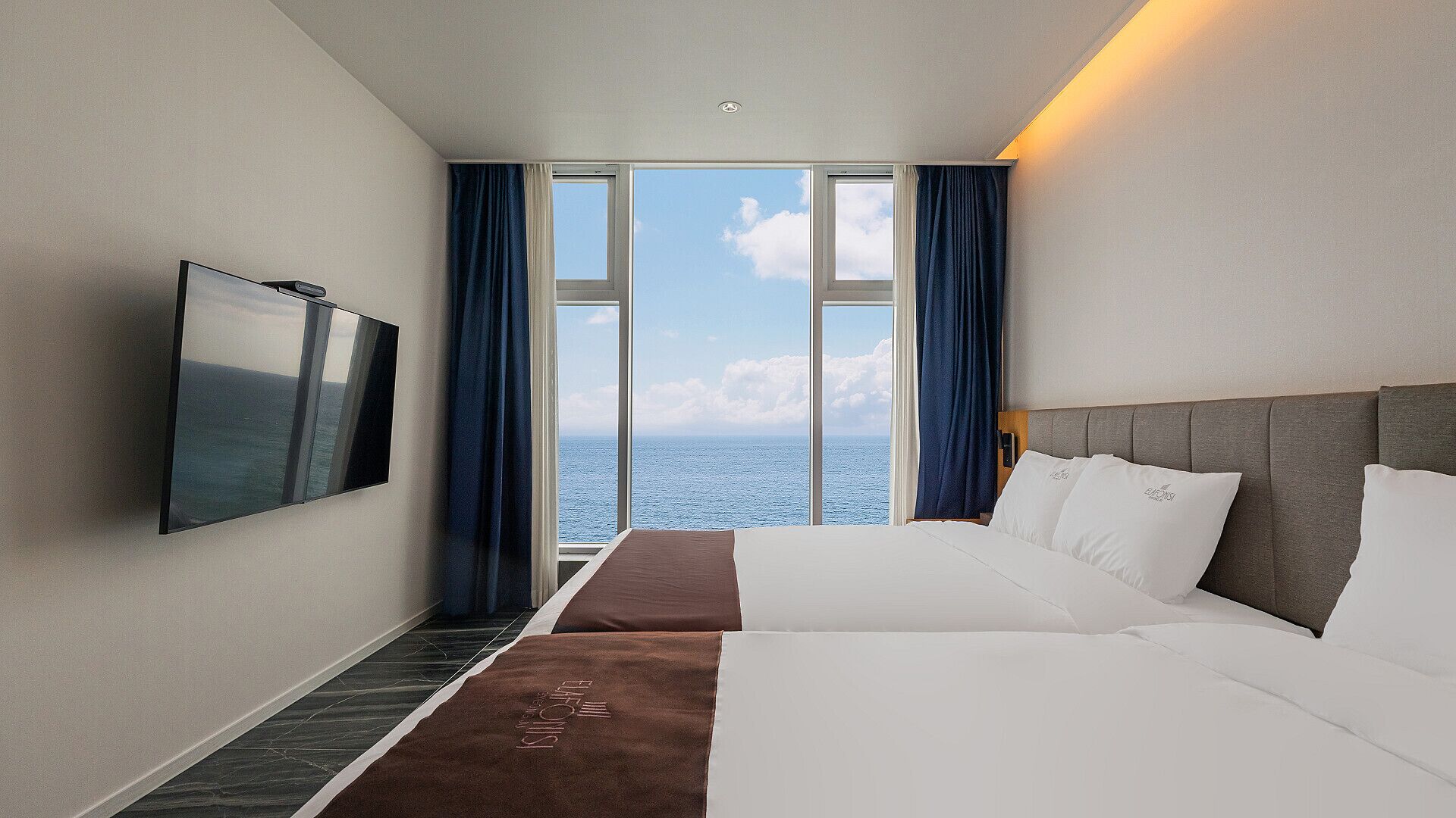 A hotel room with a large window offering a sea view, a flat-screen TV mounted on the wall, and a bed with white linens.