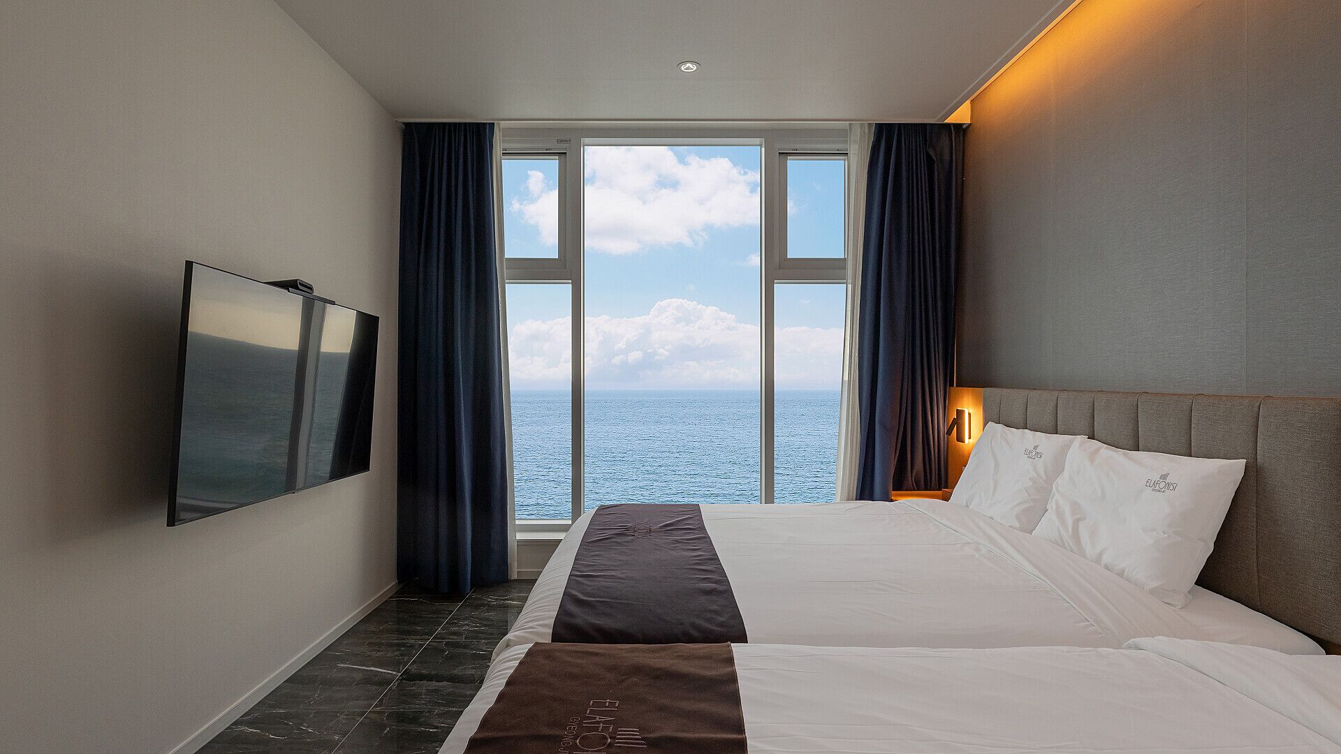 Ein Hotelzimmer mit einem großen Bett, einem an der Wand montierten Fernseher und Meerblick durch große Fenster.