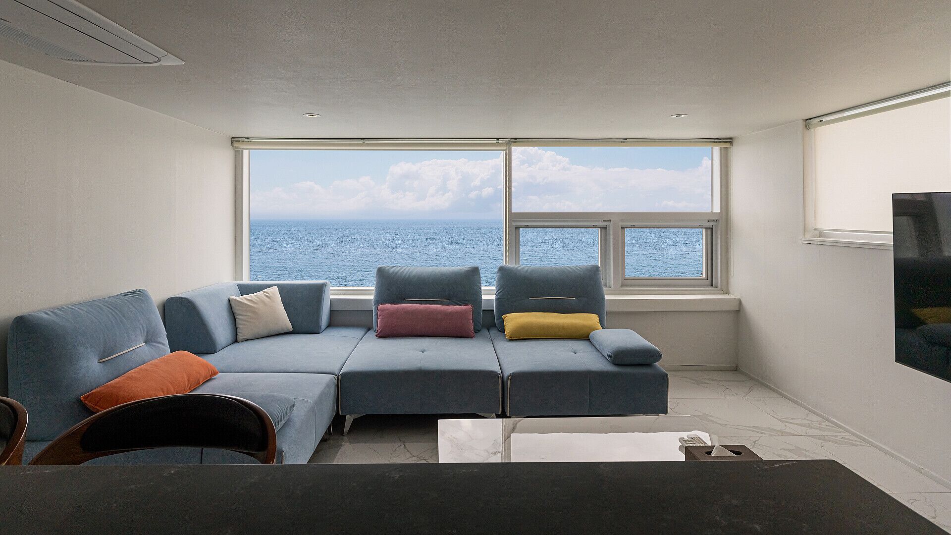 A modern living room with a large window overlooking the sea, a sofa with colorful cushions, and a flat-screen TV mounted on the wall.