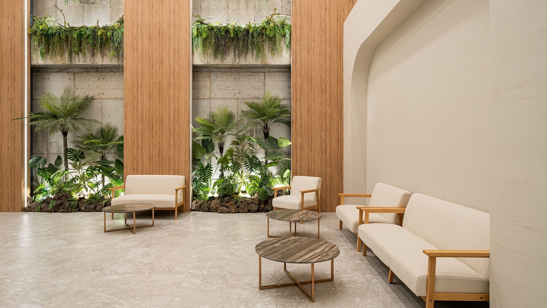 A modern indoor space with a seating area featuring a sofa and chairs, surrounded by lush greenery and a concrete wall.