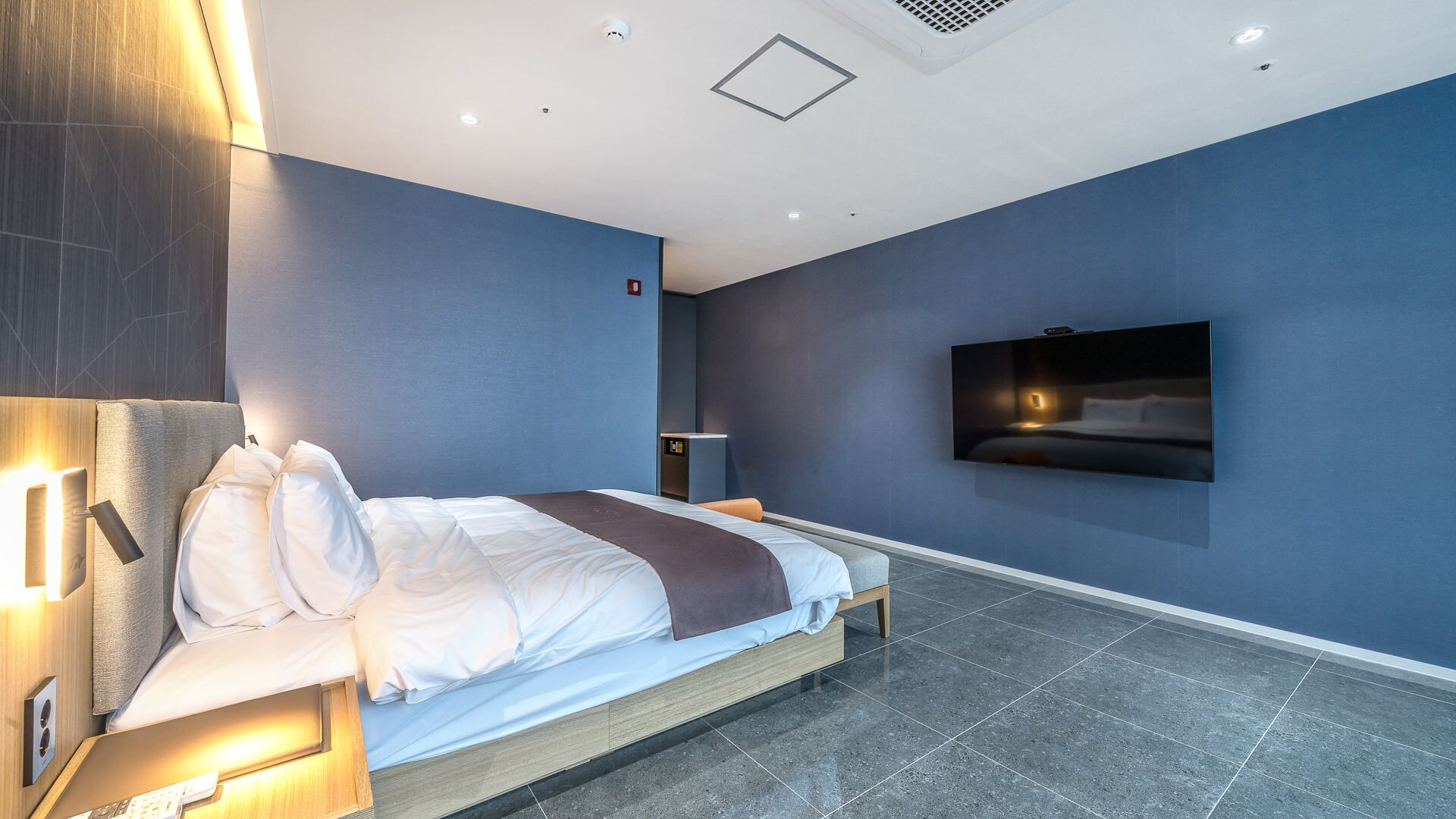 A modern hotel room with a large bed, a flat-screen TV mounted on the wall, and a small desk with a chair.