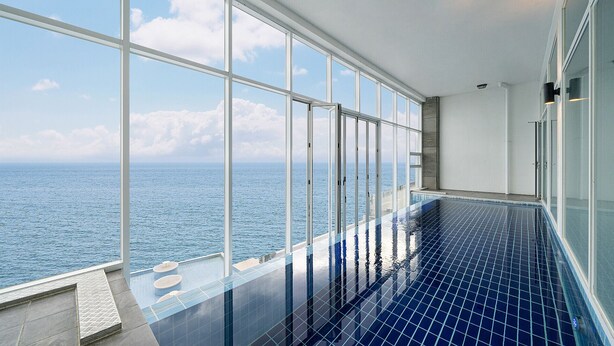 Una piscina infinita en el interior con vistas al océano, ventanas amplias y suelo de baldosas.
