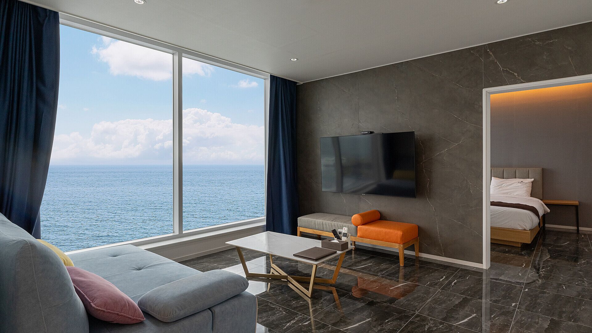 A modern hotel room with a large window offering a sea view, a flat-screen TV mounted on the wall, a comfortable seating area with a sofa and ottoman, and a separate bedroom with a bed and a nightstand.