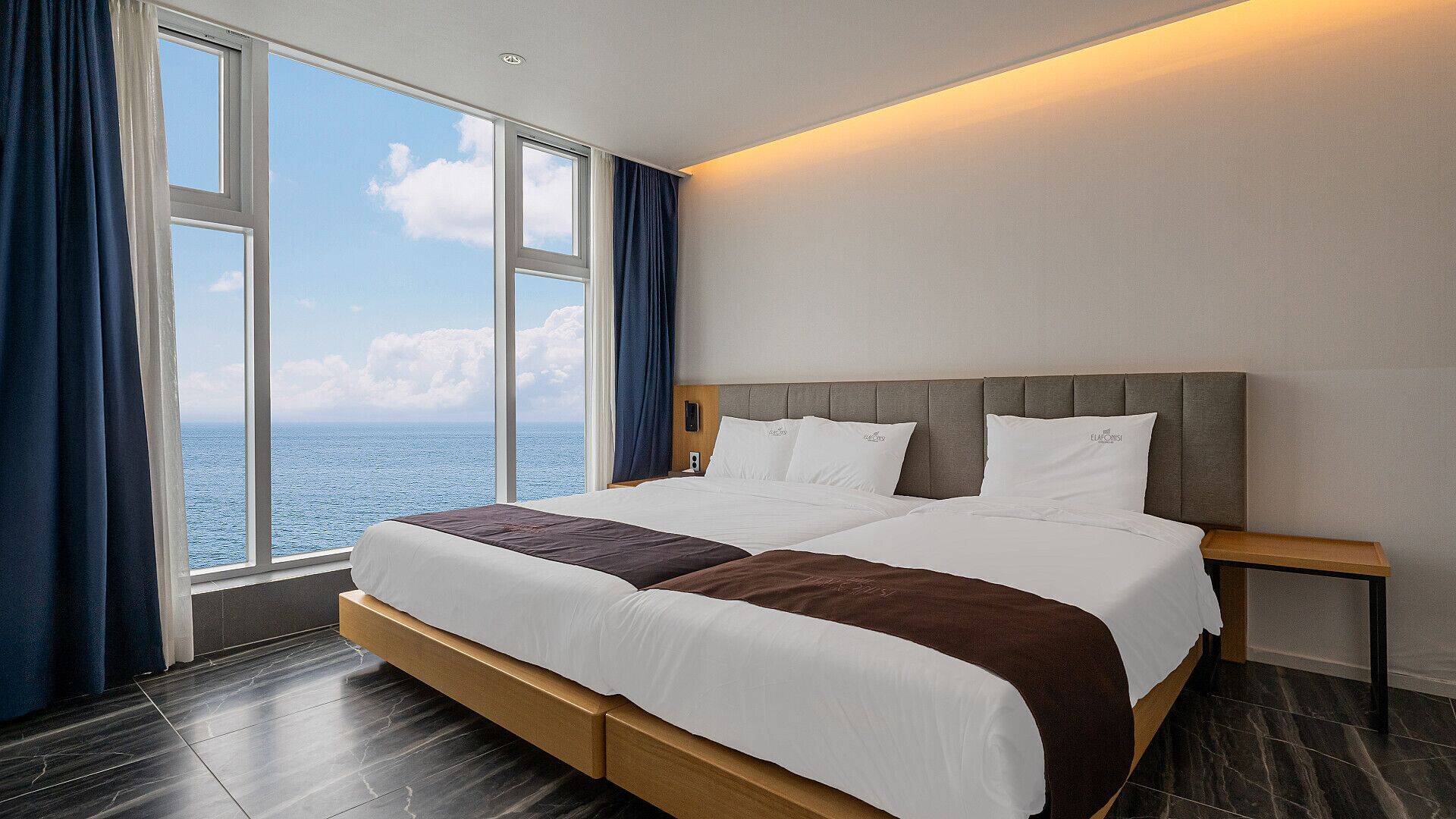 A double bed with a wooden frame and a headboard, positioned next to a large window with a view of the sea.
