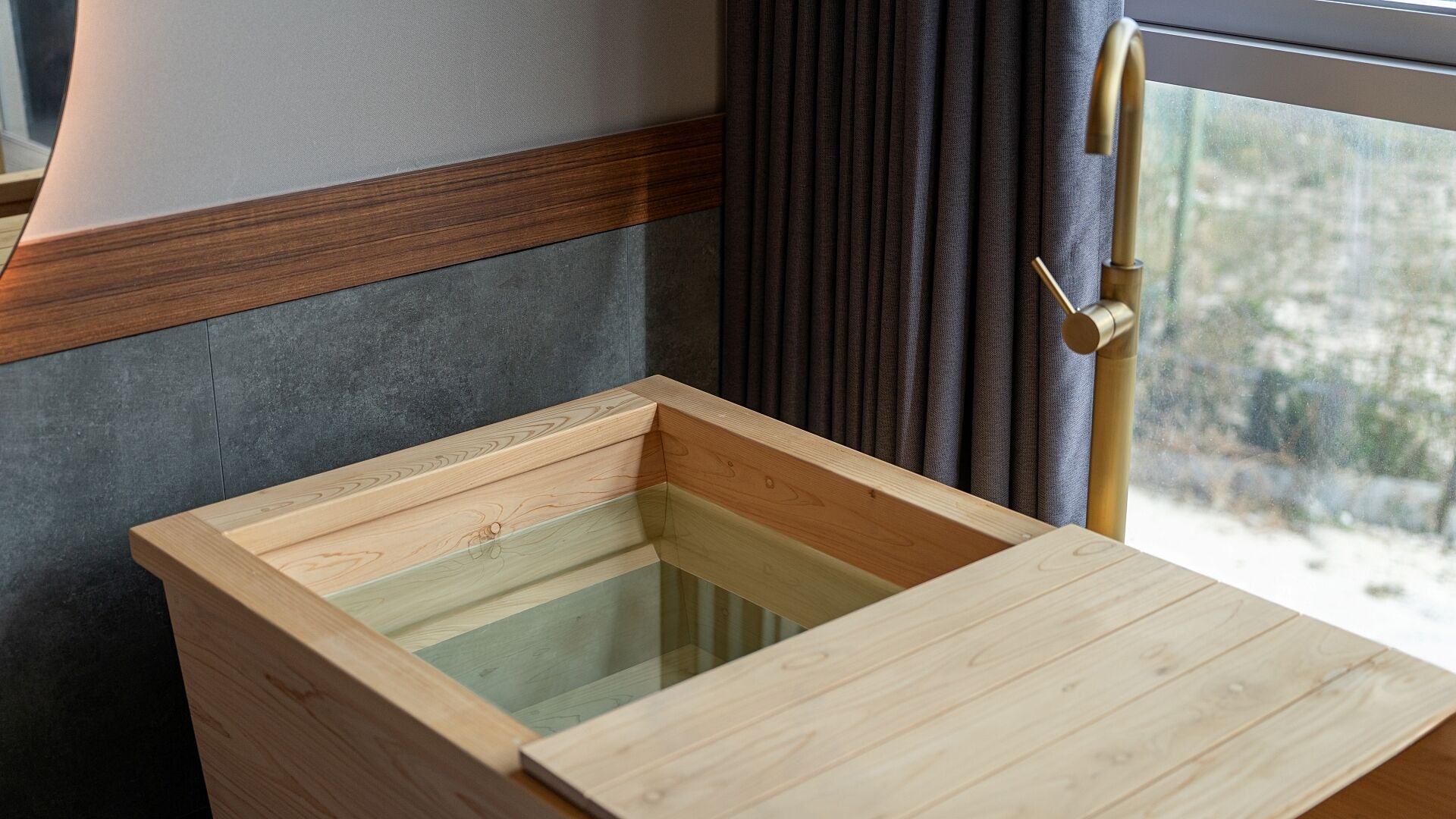 A wooden hot tub with a glass enclosure, set against a dark wall with a window and curtains.