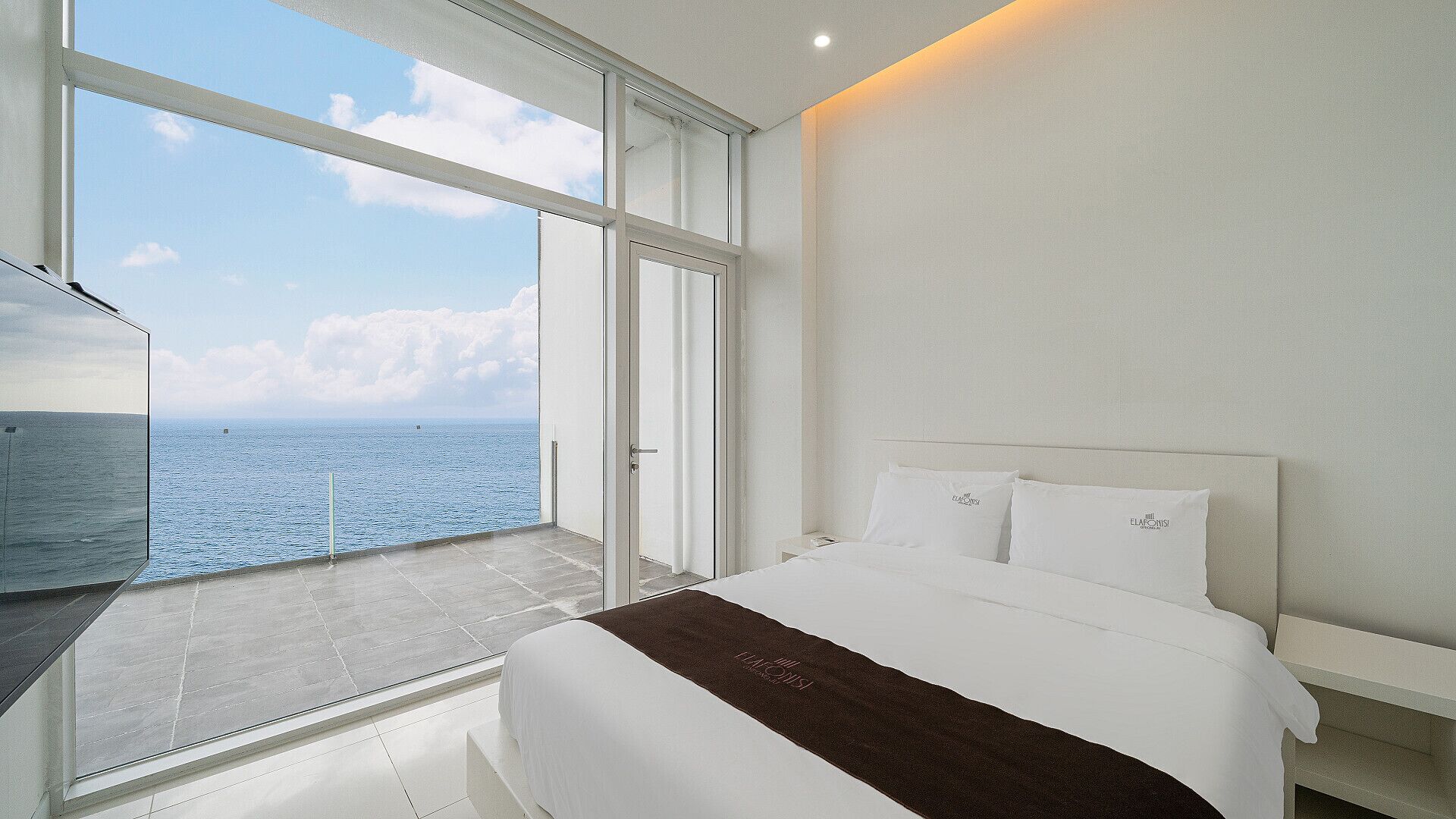 Un dormitorio moderno con un ventanal grande que da al mar, una cama con ropa de cama blanca y vistas al océano.