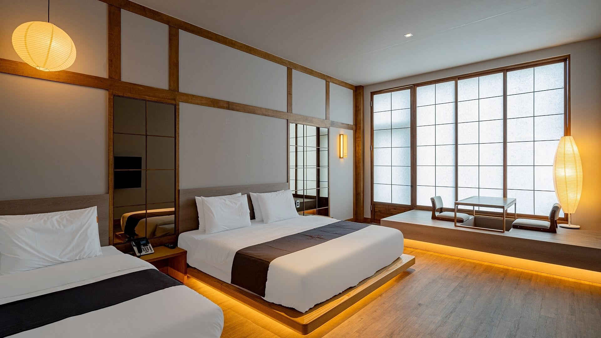 Habitación de hotel con dos camas, una zona de estar con mesa y sillas, ventanas amplias y una lámpara de estilo japonés tradicional.