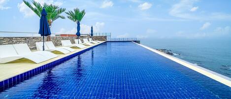 A long, narrow infinity pool with white lounge chairs and blue umbrellas, overlooking the ocean.