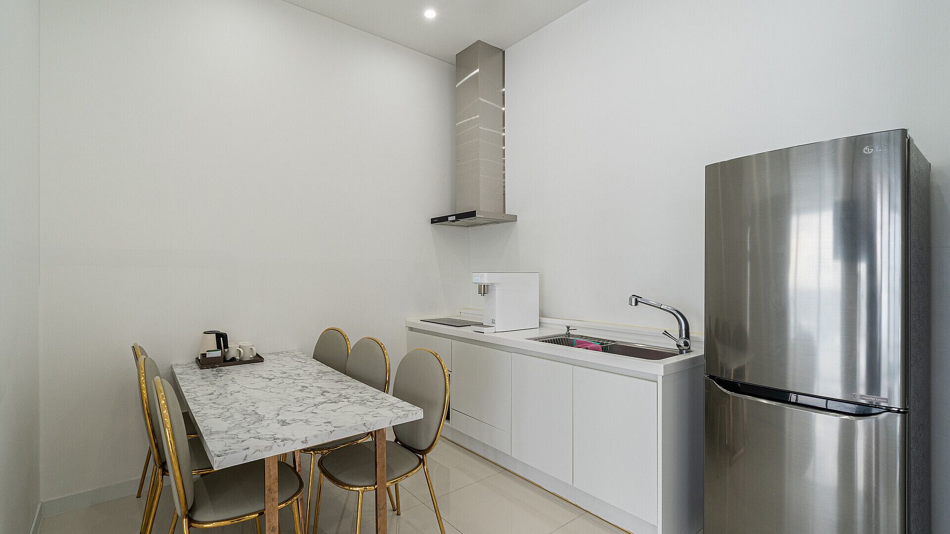 A compact kitchen with a marble-top table, four chairs, a stainless steel refrigerator, a built-in oven, and a coffee machine.