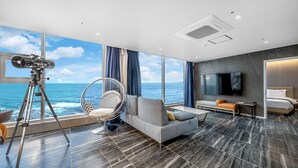 Una habitación de hotel moderna con un ventanal amplio que ofrece vistas al mar, un sofá confortable, una cama, un televisor y un telescopio sobre un trípode.