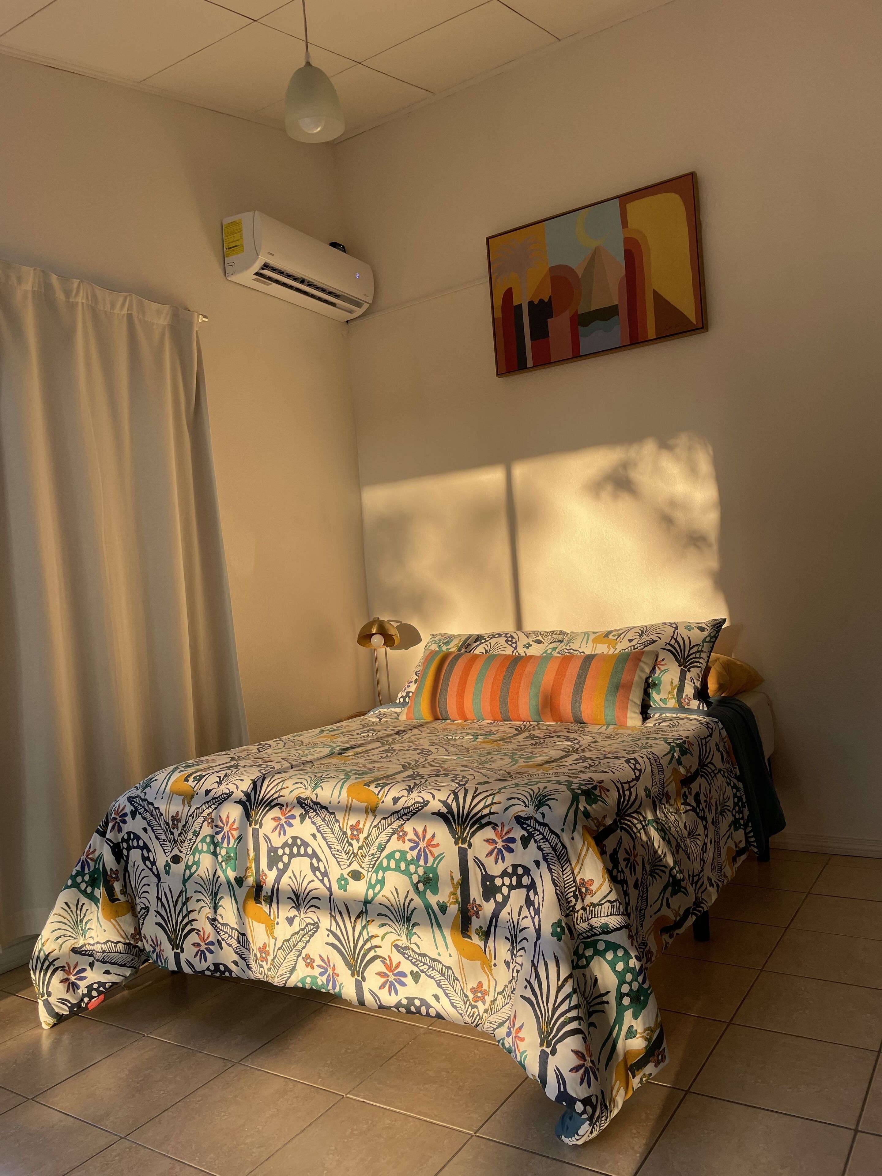 Una camera da letto con un letto, tende, un condizionatore, una lampada e un quadro appeso al muro.