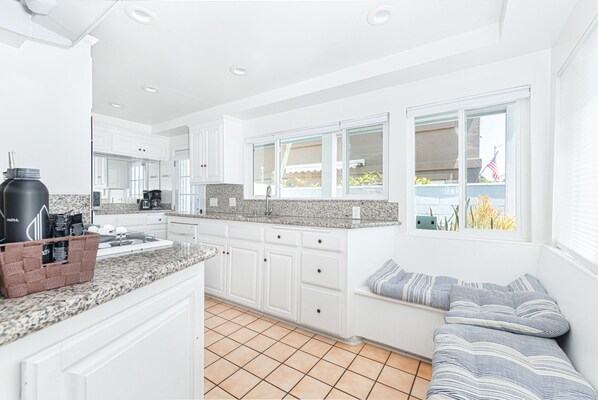 A modern kitchen with white cabinets, granite countertops, and a built-in oven. There is a window with a view of the outside, and a small seating area with a striped cushion.