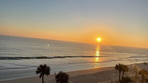 A beach with the sun setting over the ocean, palm trees, and a small building with a staircase. - A breathtaking oceanfront studio w/ a private balcony (Daytona Beach)