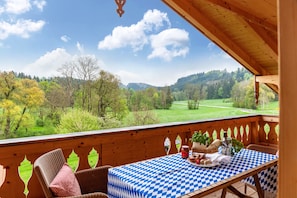 A wooden deck with a checkered tablecloth, wicker chairs, and a scenic view of a forested valley.