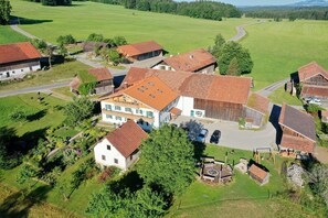 An aerial view of a rural village with traditional houses, a central building with a sloped roof, and surrounding green fields.