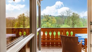 A wooden deck with a view of a forested landscape, a wicker chair, and a table with a checkered tablecloth. - Apartment 'Xaver' with Shared Garden, Balcony, and Wi-Fi (Rottenbuch)