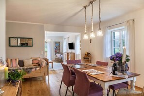 A dining area with a wooden table, chairs, and a chandelier. There is a sofa with cushions and a television in the background. - Apartment "Lina" with Shared Garden, Balcony, and Wi-Fi (Rottenbuch)