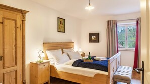 Una cama de madera con cabecera, una mesita de noche con lámpara, un banco y una ventana con cortinas.