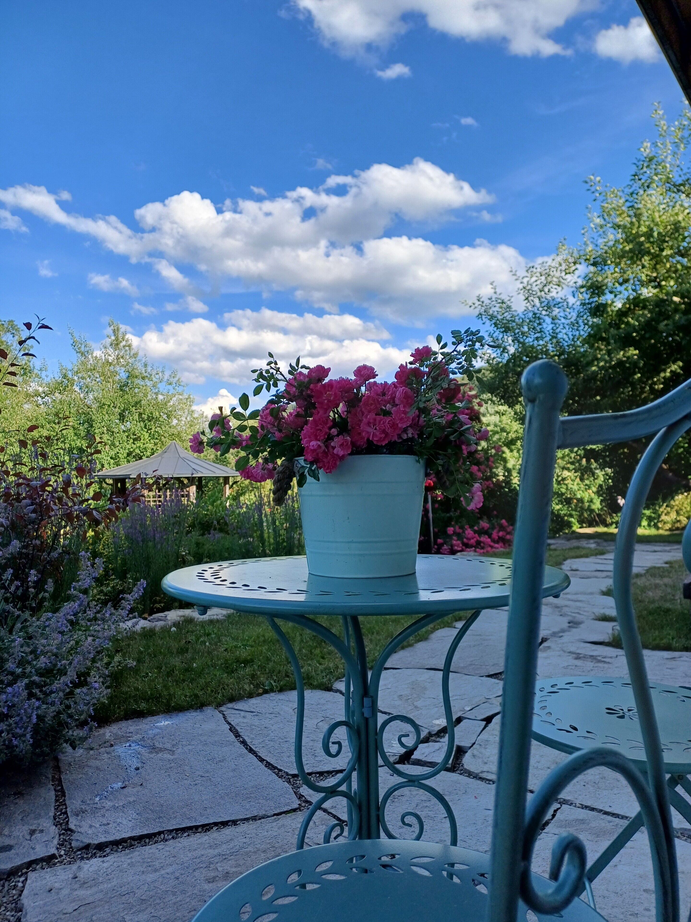 A garden patio with a metal table and chair, a potted plant with pink flowers, and a gazebo in the background.