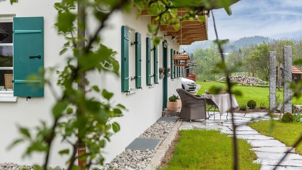 A house with a white exterior, green shutters, a patio with a table and chairs, and a view of a mountainous landscape. - Apartment 'Lisbeth' with Shared Garden, Balcony, and Wi-Fi (Rottenbuch)