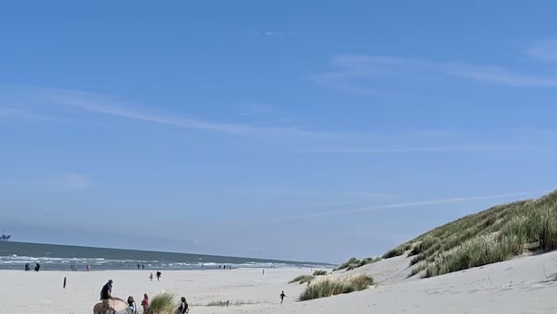Ein Strand mit Menschen, die laufen und stehen, einem klaren Himmel und Dünen im Hintergrund.