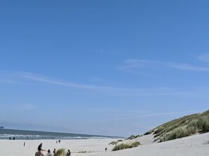 Une plage où l’on voit des gens se promener et se tenir debout, un ciel dégagé et des dunes en arrière-plan.