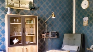 Ein Zimmer mit blauen Tapeten, einem Holzkabinett mit Glastüren, einem blauen Sessel, einer Stehlampe und einer Wanduhr.
