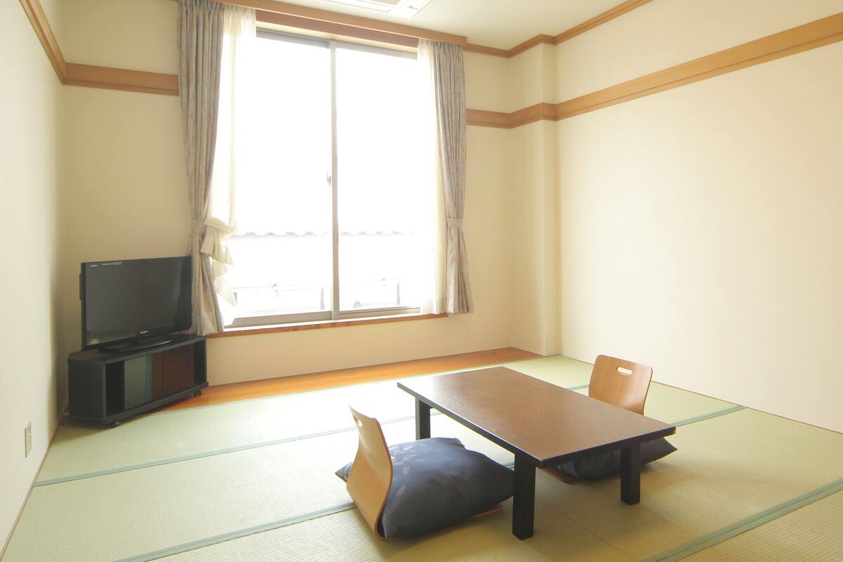Ein Zimmer mit Tatami-Böden, einem niedrigen Tisch, einem Stuhl, einem Fernseher und einem großen Fenster mit Vorhängen.