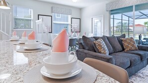 Un salon moderne avec un canapé gris, un coin repas avec une table blanche et une cuisine dotée d’un plan de travail en granit.