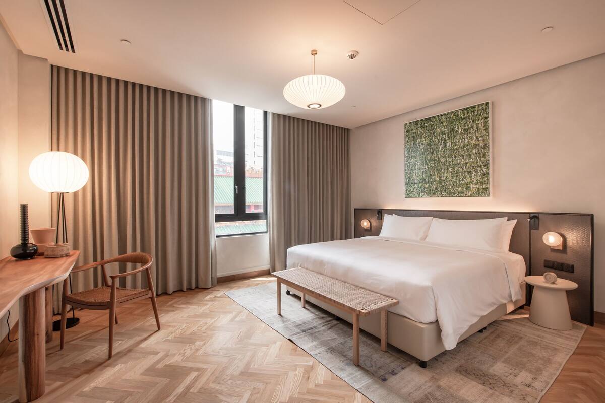 A modern hotel room with a large bed, a desk with a chair, a bench, a bedside table, and a wall-mounted artwork.