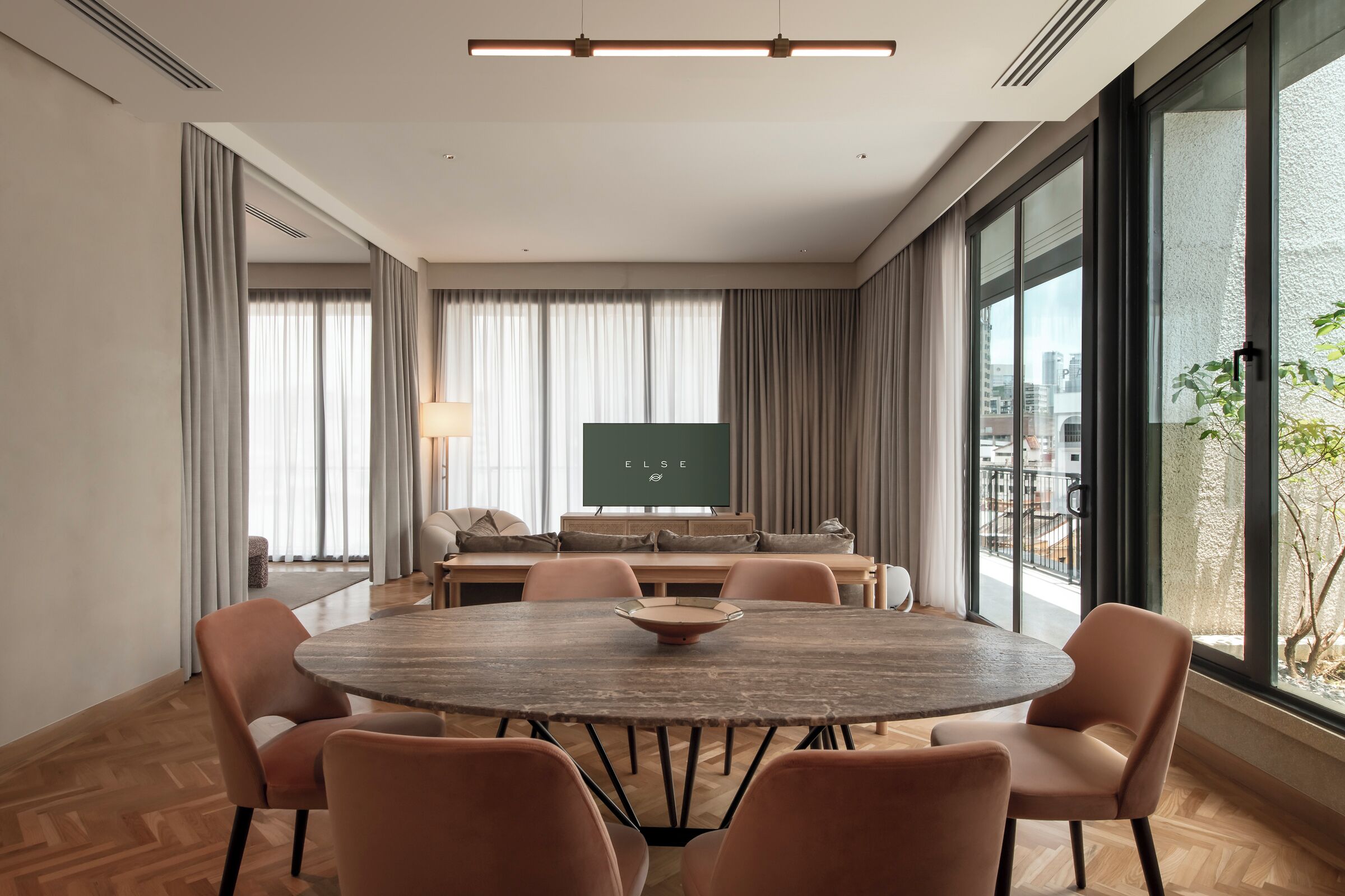 A modern conference room with a round wooden table, cushioned chairs, a large window with a view of buildings, and a projection screen.
