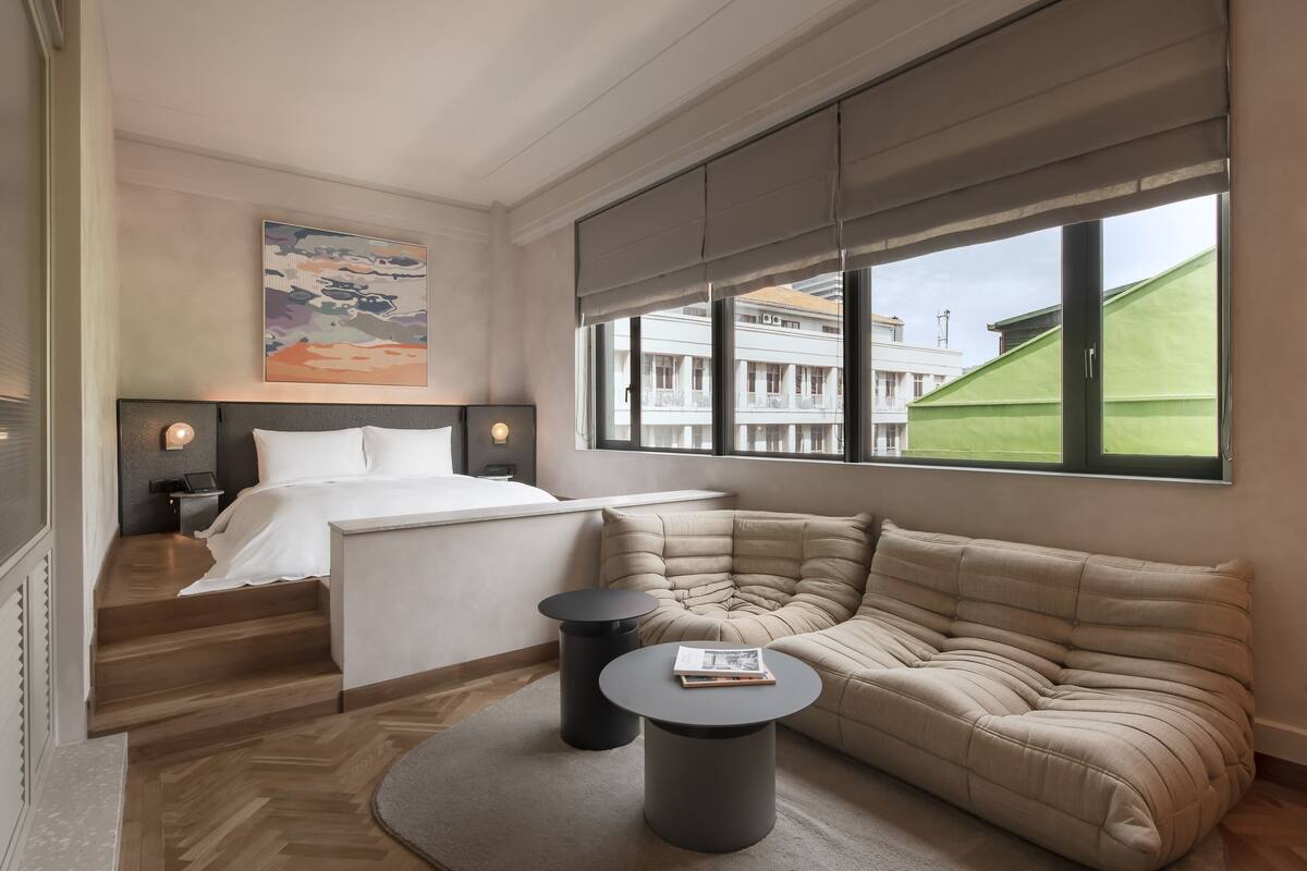 A modern hotel room with a bed, a sofa, a small table, and a large window offering a view of buildings.
