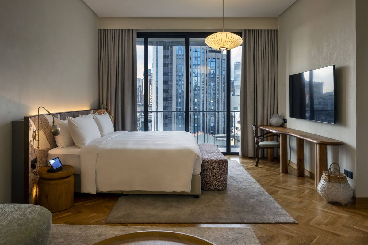 A modern hotel room with a large bed, a desk, a TV, and a view of the city through large windows.