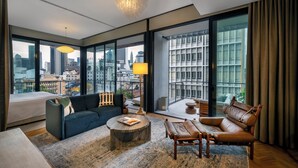 A modern hotel room with a large bed, a dark blue sofa, a leather armchair, a round coffee table, and a floor lamp.