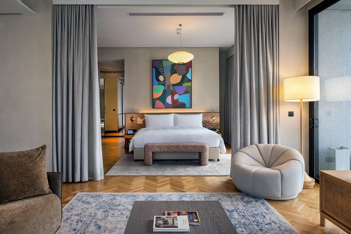 A modern hotel room with a large bed, a sitting area with a chair, a coffee table with books, a painting above the bed, and a view through the window.