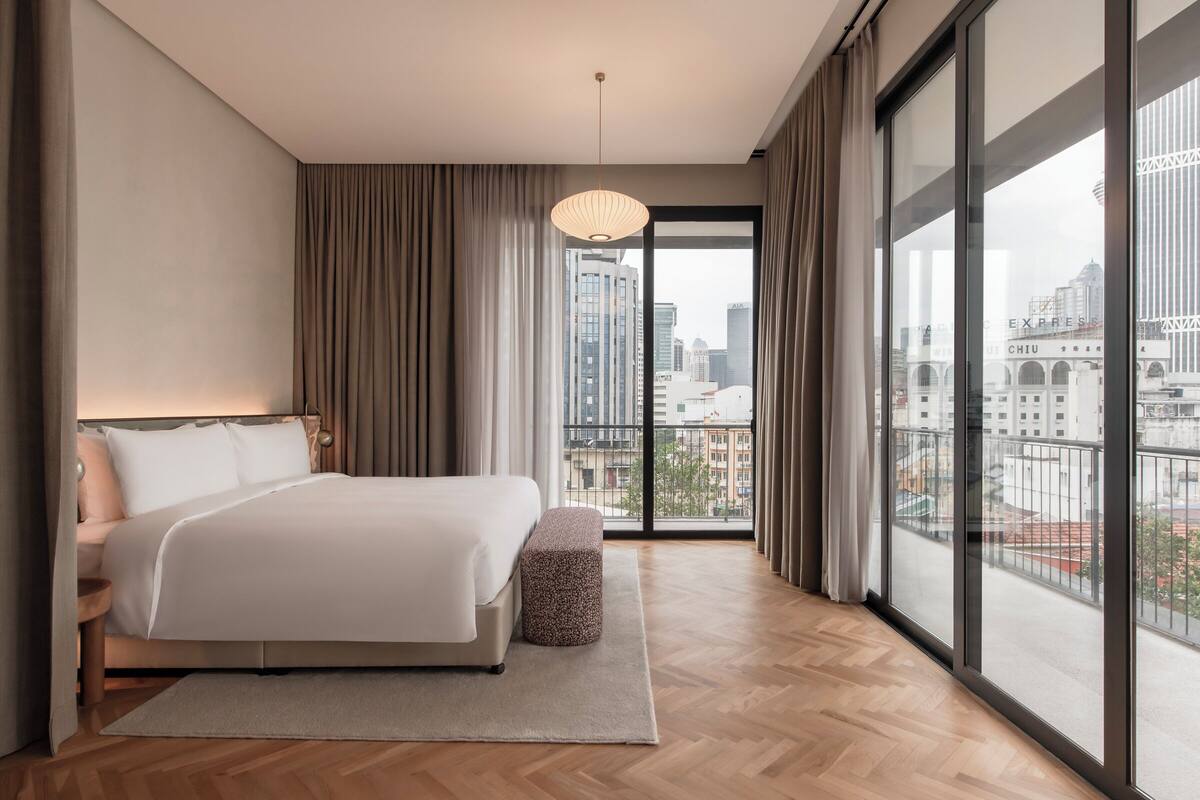 A modern hotel room with a large bed, a small ottoman, and a view of the city through floor-to-ceiling windows.