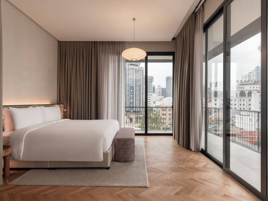 Una habitación de hotel moderna con una cama grande, un pequeño reposapiés y vistas de la ciudad a través de ventanales de gran altura.