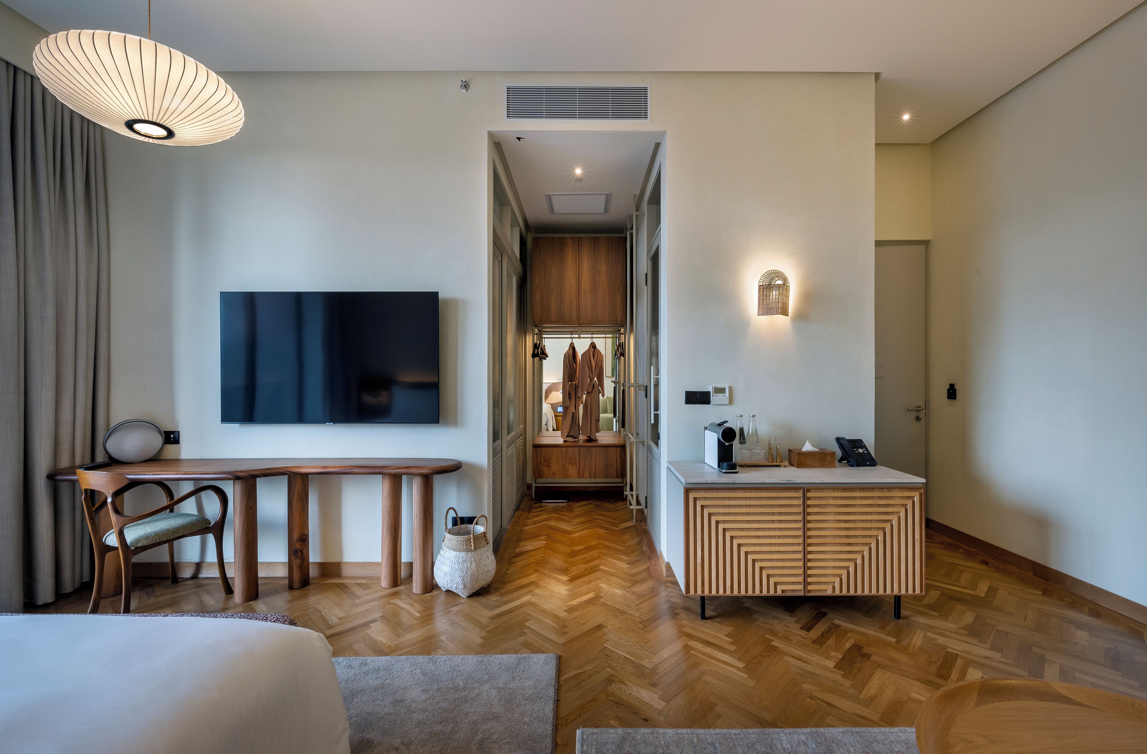 A modern hotel room with a wooden floor, a large bed, a desk with a chair, a TV, a wardrobe, and a small kitchenette.