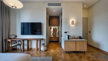 A modern hotel room with a wooden floor, a large bed, a desk with a chair, a TV, a wardrobe, and a small kitchenette.