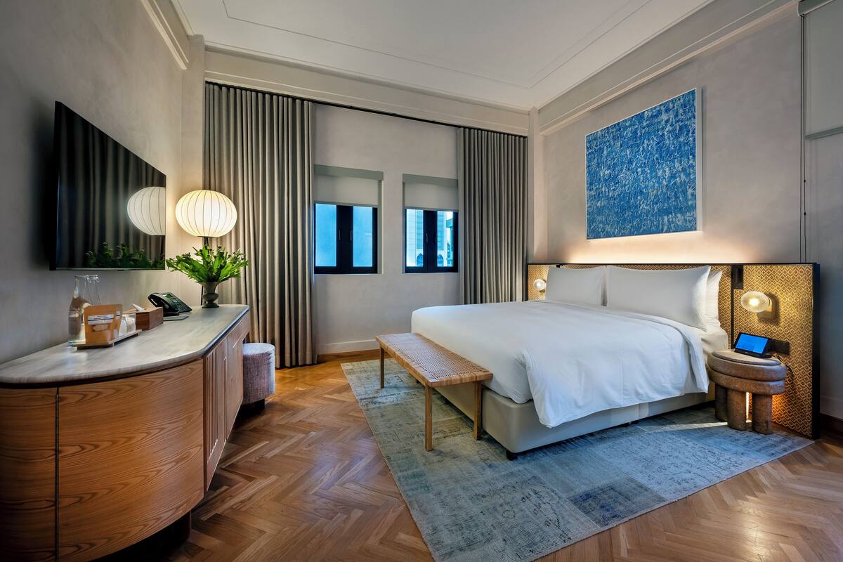 A modern hotel room with a large bed, a desk with a lamp, a small table, and a nightstand with a laptop.