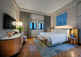 A modern hotel room with a large bed, a desk with a lamp, a small table, and a nightstand with a laptop.