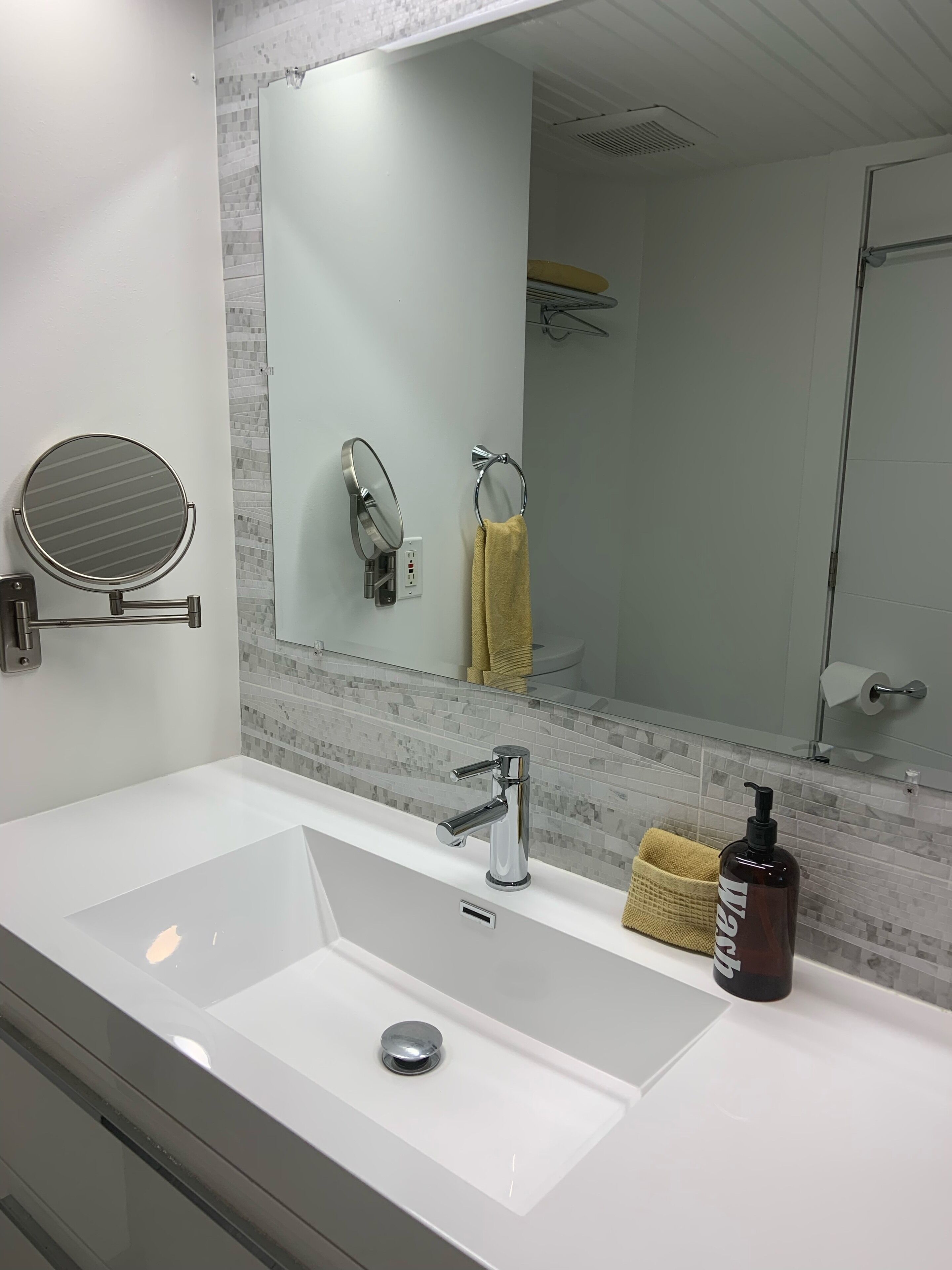 A modern bathroom with a large mirror, a round mirror, a chrome faucet, and a beige towel on the counter.