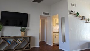 A living room with a flat-screen TV, a wooden console table with books, and a plant. There is a kitchen visible through an open doorway. - Tucson Pool House, a vacation home with easy access to all Tucson offers (Tucson)