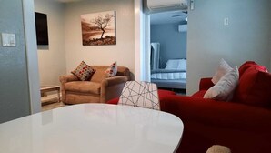 Un salon avec une table blanche, un fauteuil marron et un canapé rouge. Il y a un tableau au mur et un lit à l’arrière-plan.