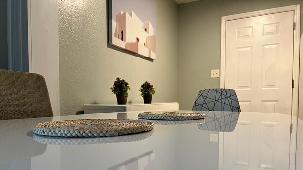 Une table à manger avec des sets de table en fibres tressées, une chaise à motifs géométriques, un tableau accroché au mur et une porte visible au fond.