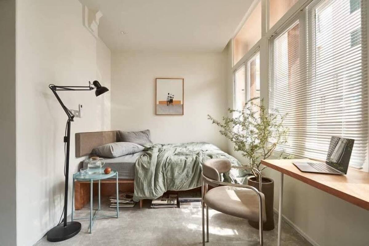 A bedroom with a bed, a desk with a laptop, a chair, a floor lamp, and a window with blinds.